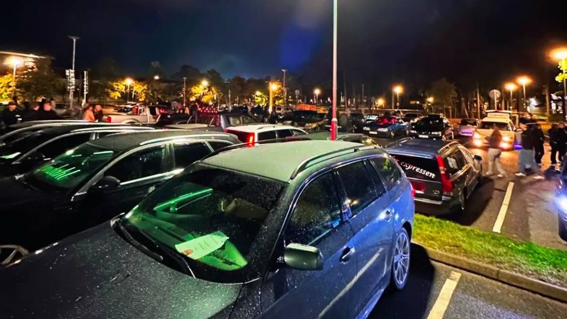 Parkerade bilar och människor samlade på en upplyst parkeringsplats kvällstid.