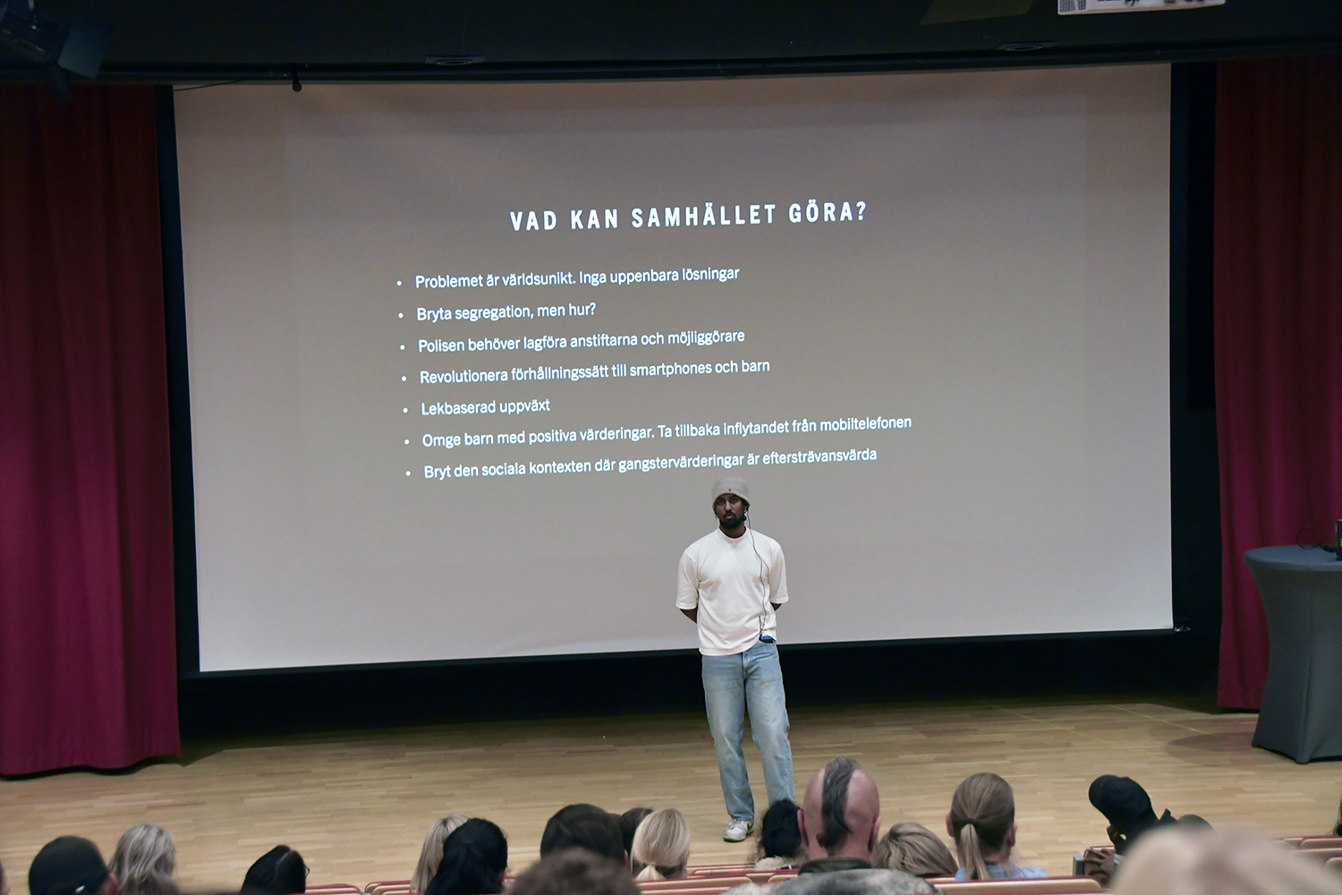 Föreläsaren Luay Mohageb håller en presentation på scen framför en stor skärm med text om förslag på samhällsåtgärder.