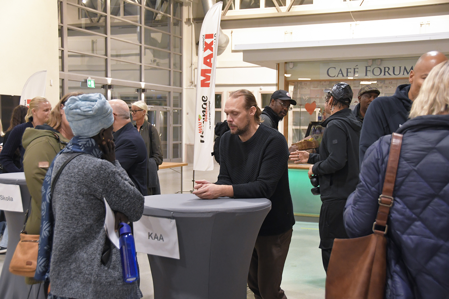 Deltagare samtalar vid höga bord. En skylt med texten 'KAA' och en banner från Maxi Stormarknad Haninge syns i bakgrunden, som sponsor av fikat från kvällen.