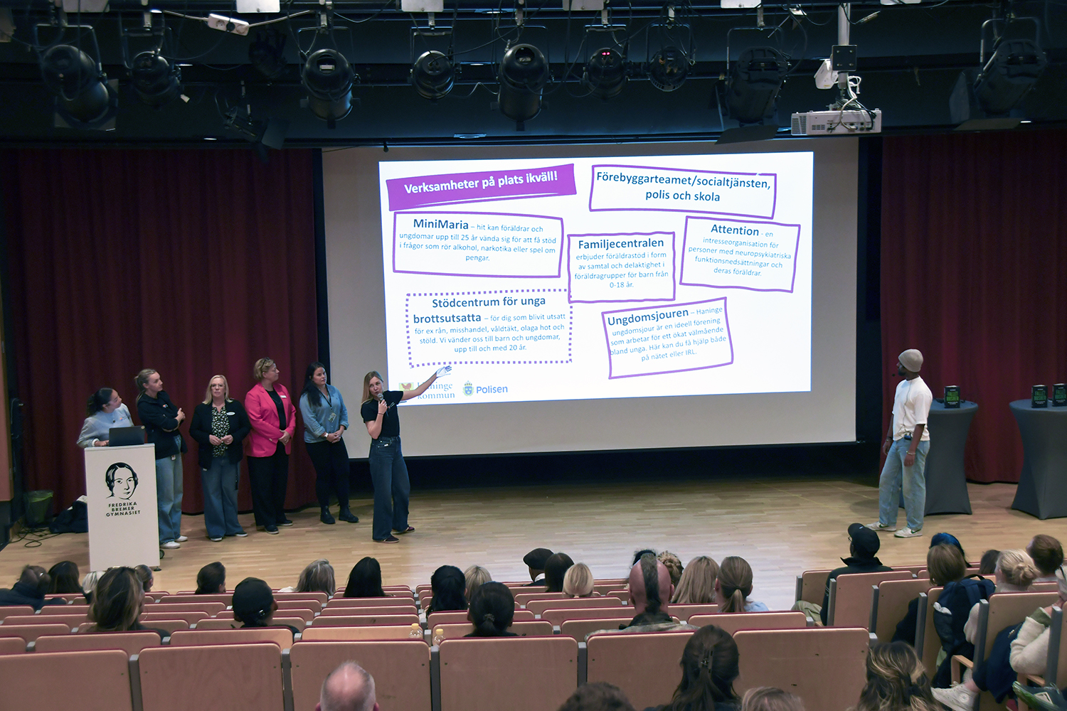 Presentation på scen med flera personer och en skärm som visar vilka lokala stödverksamheter som är på plats, inför en sittande publik.