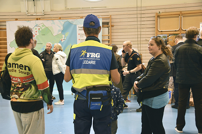 Personer samlade vid ett informationsbord under invigningen av trygghetspunkter. En person bär väst med texten 'FRG LEDARE' och en stor karta med trygghetspunkterna syns i bakgrunden.